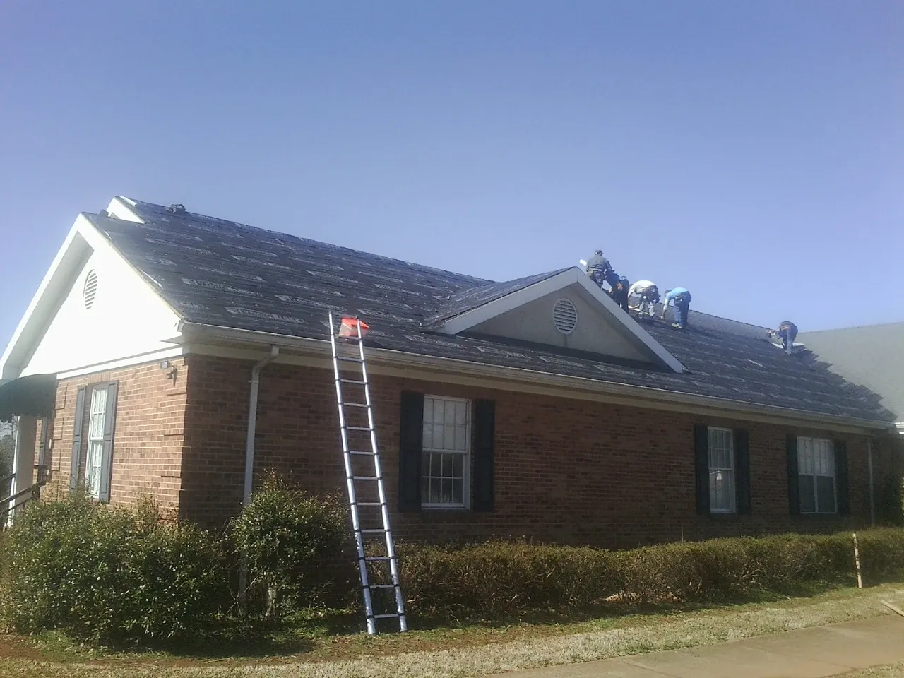 Crew tearing shingles off a brick ranch roof with a ladder set — Metal Roof Repair work by our crew in Lyndonville